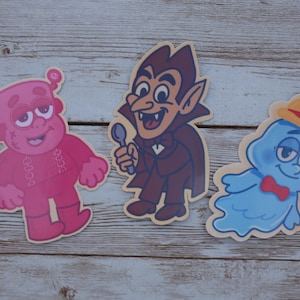 Puede incluir: Tres pegatinas de personajes de dibujos animados. Una figura rosa parecida a Frankenstein, un vampiro sosteniendo una cuchara y un fantasma azul con pajarita y sombrero. Las pegatinas están sobre una superficie de madera.