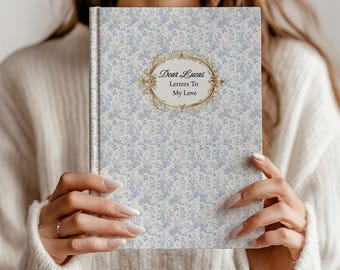 May include: A hardcover journal with a light blue floral pattern. The cover features an ornate gold frame with the text "Dear Lucas Letters To My Love". The journal is held by a person wearing a white sweater.