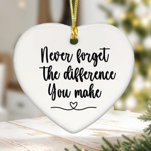 May include: White heart-shaped ornament with the text "Never forget the difference You make" in black script. A small heart is drawn below the text. The ornament hangs from a gold cord.