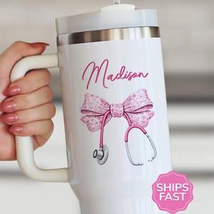 May include: White insulated tumbler with a handle, featuring a pink bow, stethoscope, and the name "Madison" in cursive. The design is on a white background. The tumbler has a silver rim and a white lid.