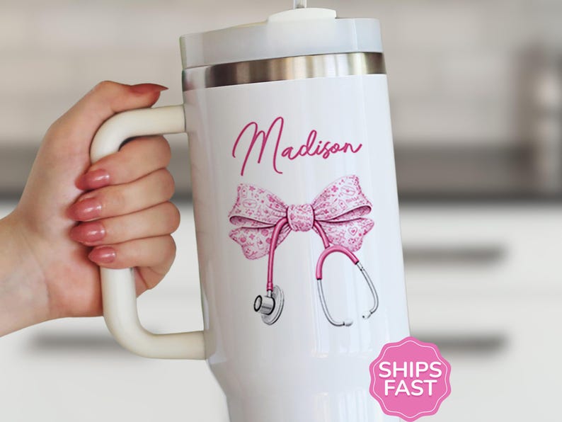 May include: White insulated tumbler with a handle, featuring a pink bow, stethoscope, and the name "Madison" in cursive. The design is on a white background. The tumbler has a silver rim and a white lid.