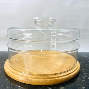 May include: A clear glass cloche with a round wooden base. The glass dome has a small handle on top and sits atop a light-colored wooden base. The cloche is designed to protect and display items.