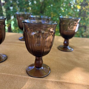 May include: A collection of brown-tinted glass goblets with a decorative pattern. The goblets have a rounded bowl shape, a short stem, and a circular base. The glasses are arranged on a tan surface, with a blurred green background.