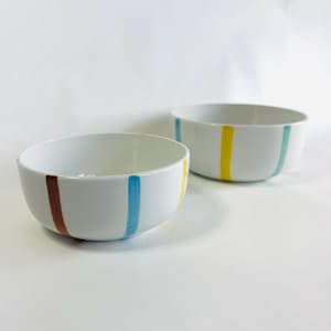 May include: Two white ceramic bowls with colorful vertical stripes. One bowl has brown, blue, and yellow stripes, while the other has blue, yellow, and teal stripes. The bowls are round and have a simple, modern design.