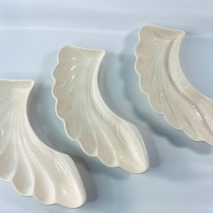 May include: Three off-white, wing-shaped ceramic dishes are arranged on a light blue surface. Each dish has a scalloped edge and a textured, feather-like design. The dishes are likely for decorative or serving purposes.