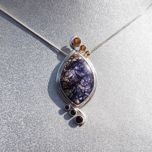 May include: A silver pendant necklace featuring a teardrop-shaped stone with a purple and white pattern. The pendant is accented with small, round, amber and purple stones. The necklace hangs on a thin silver chain.