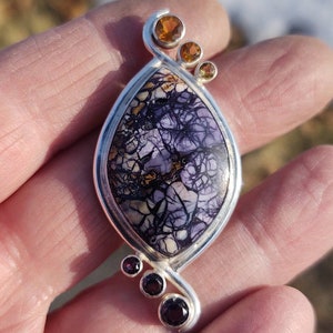 May include: A handcrafted pendant with a silver setting. The focal point is a large, oval-shaped stone with a purple, cream, and black pattern. Accented with three orange and three purple gemstones. A unique piece of jewelry.