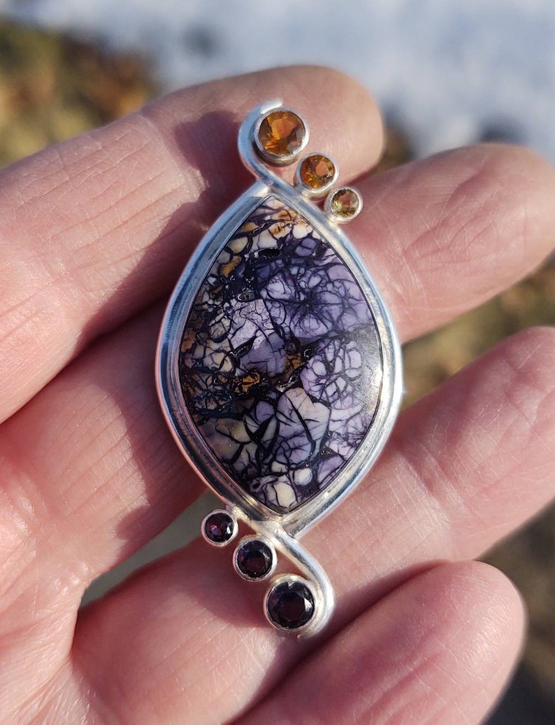 May include: A handcrafted pendant with a silver setting. The focal point is a large, oval-shaped stone with a purple, cream, and black pattern. Accented with three orange and three purple gemstones. A unique piece of jewelry.