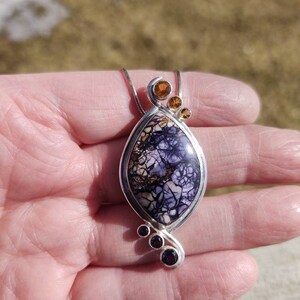 May include: A silver pendant necklace featuring a teardrop-shaped stone with a purple and cream pattern. The pendant is accented with three small orange-brown stones at the top and three smaller purple stones at the bottom.