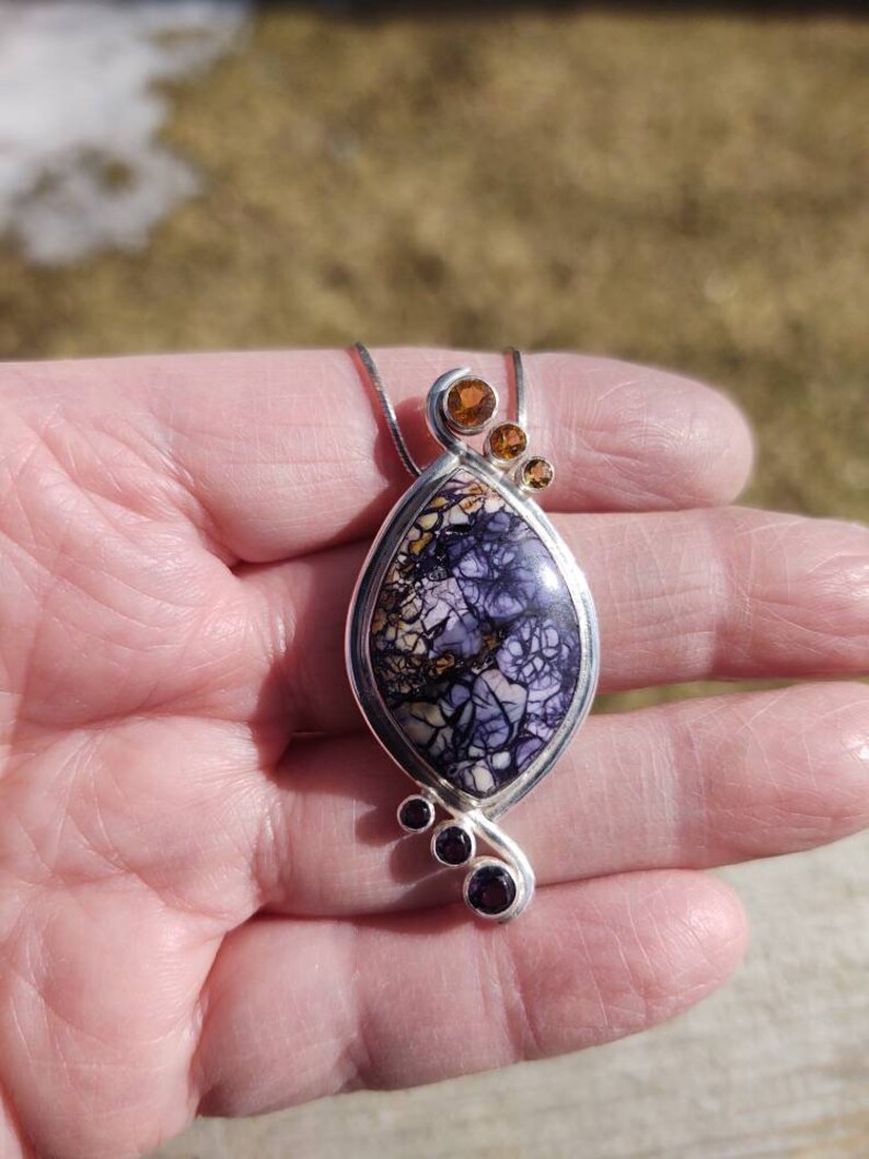 May include: A silver pendant necklace featuring a teardrop-shaped stone with a purple and cream pattern. The pendant is accented with three small orange-brown stones at the top and three smaller purple stones at the bottom.