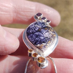 May include: A silver pendant with a large, oval-shaped stone featuring a striking pattern of purple and white veining. The pendant is accented with three small, round, orange-yellow stones and three small purple stones. The pendant hangs from a silver chain.