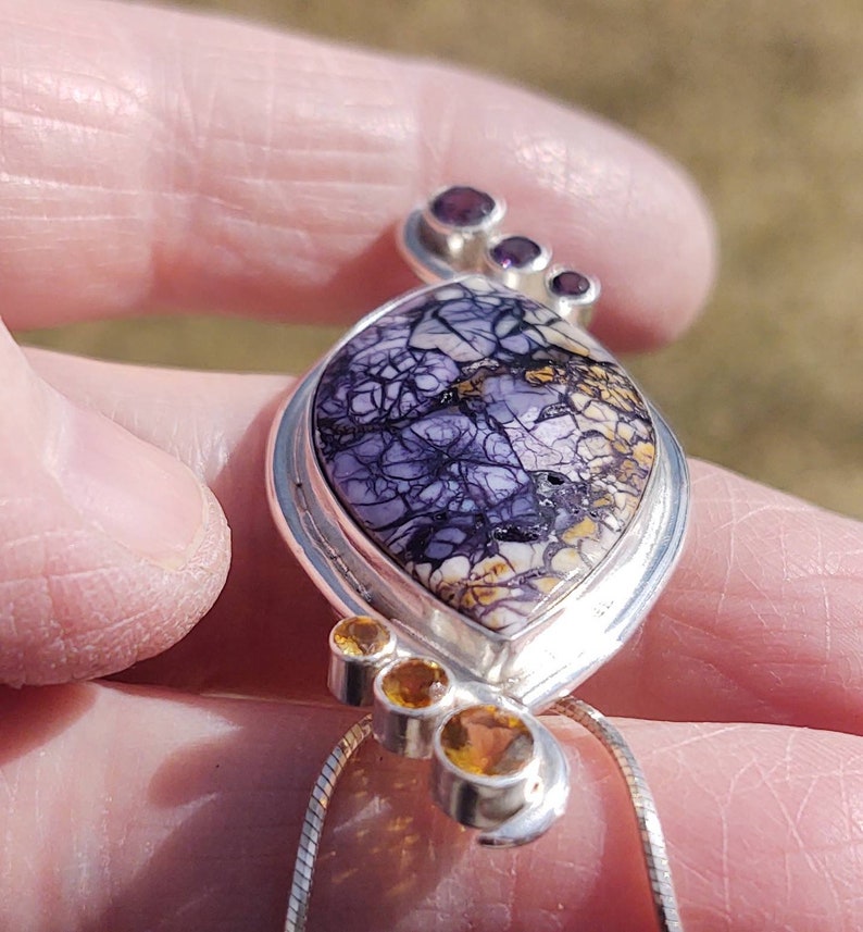 May include: A silver pendant with a large, oval-shaped stone featuring a striking pattern of purple and white veining. The pendant is accented with three small, round, orange-yellow stones and three small purple stones. The pendant hangs from a silver chain.