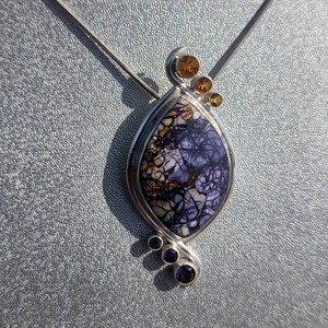 May include: A silver pendant necklace featuring a teardrop-shaped stone with a unique pattern of purple, white, and brown. The pendant is accented with small orange and purple gemstones. The necklace is displayed on a silver chain.