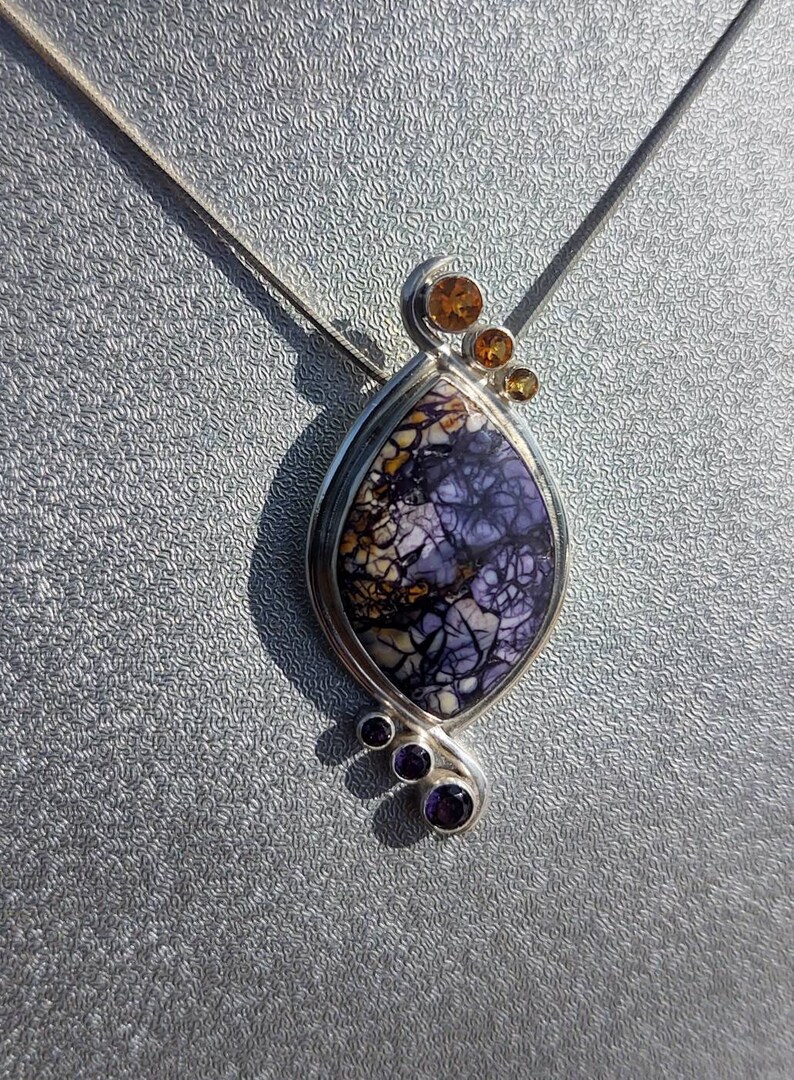 May include: A silver pendant necklace featuring a teardrop-shaped stone with a unique pattern of purple, white, and brown. The pendant is accented with small orange and purple gemstones. The necklace is displayed on a silver chain.