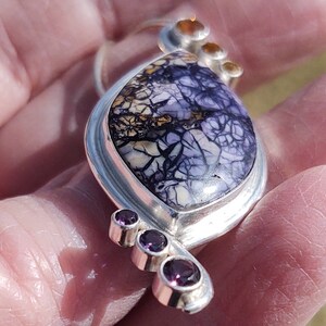 May include: A handcrafted silver pendant featuring a large, oval-shaped stone with a striking purple and white pattern. Accented with three small, round, purple gemstones and three smaller orange-brown stones. The pendant is held in a hand.