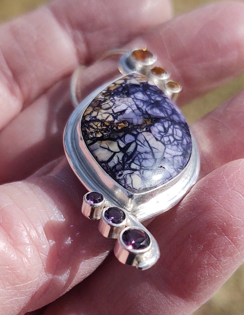 May include: A handcrafted silver pendant featuring a large, oval-shaped stone with a striking purple and white pattern. Accented with three small, round, purple gemstones and three smaller orange-brown stones. The pendant is held in a hand.
