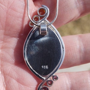 May include: Silver pendant necklace with an oval-shaped, dark-colored stone. The pendant has a decorative silver swirl design with small orange-colored accents. The necklace chain is a thin, silver snake chain. The pendant is stamped with the number "935".