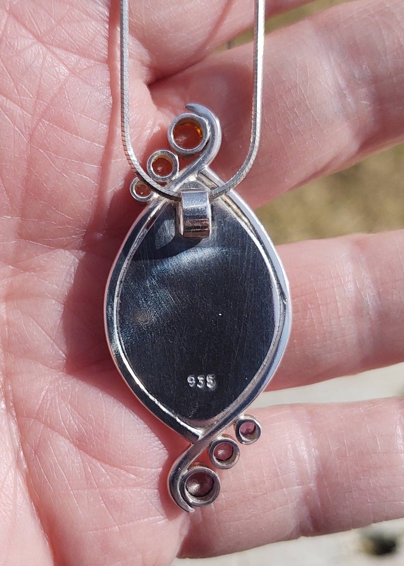 May include: Silver pendant necklace with an oval-shaped, dark-colored stone. The pendant has a decorative silver swirl design with small orange-colored accents. The necklace chain is a thin, silver snake chain. The pendant is stamped with the number "935".