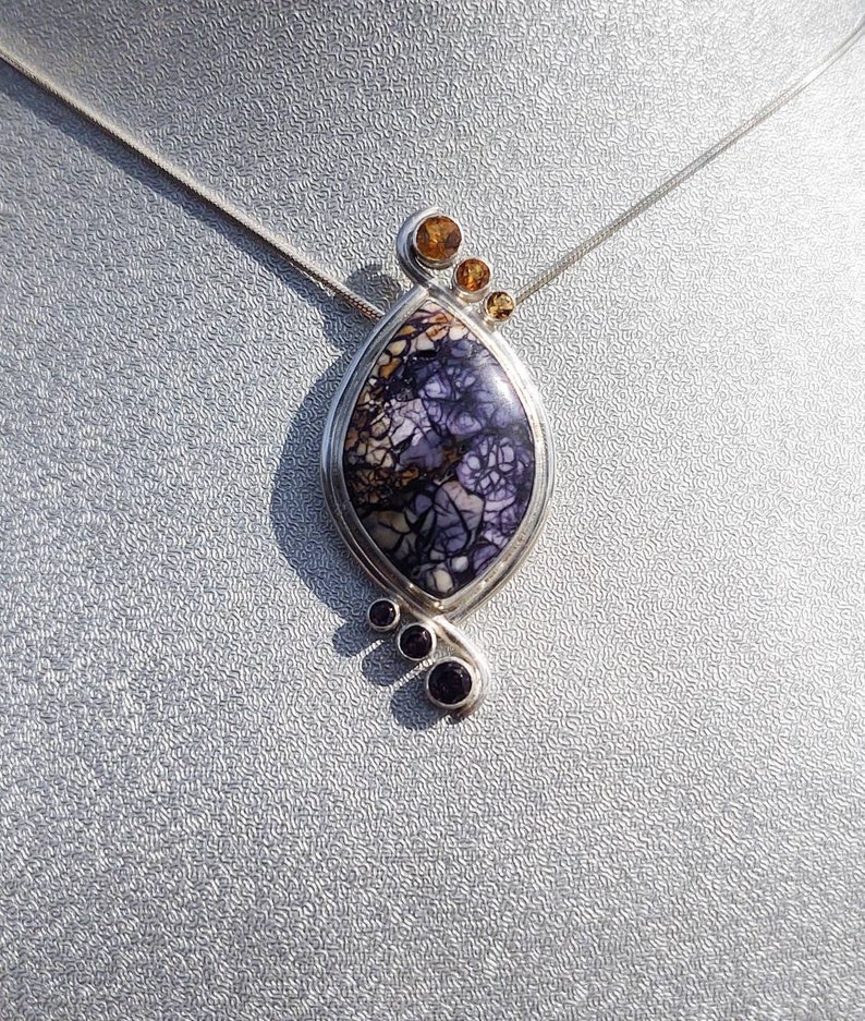 May include: A silver pendant necklace featuring a teardrop-shaped stone with a purple and white pattern. The pendant is accented with small, round, amber and purple stones. The necklace hangs on a thin silver chain.