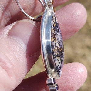 May include: A silver pendant necklace featuring a teardrop-shaped stone with a unique pattern of purple, white, and yellow. The pendant is accented with small, round, purple and yellow gemstones. The necklace is held by a silver chain.