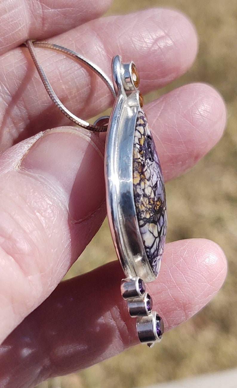 May include: A silver pendant necklace featuring a teardrop-shaped stone with a unique pattern of purple, white, and yellow. The pendant is accented with small, round, purple and yellow gemstones. The necklace is held by a silver chain.