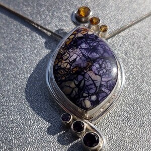 May include: A silver pendant necklace featuring a large, teardrop-shaped stone with a unique purple and brown pattern. The pendant is accented with three small, round amber-colored stones and three smaller purple stones.