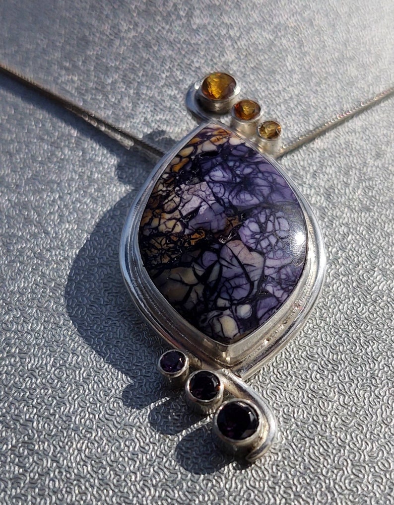 May include: A silver pendant necklace featuring a large, teardrop-shaped stone with a unique purple and brown pattern. The pendant is accented with three small, round amber-colored stones and three smaller purple stones.