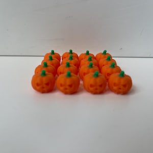 Può includere: Una collezione di piccole decorazioni a forma di zucca arancione con facce intagliate e steli verdi. Queste mini zucche sono disposte su una superficie bianca, perfette per l'artigianato e le decorazioni a tema Halloween o autunno.