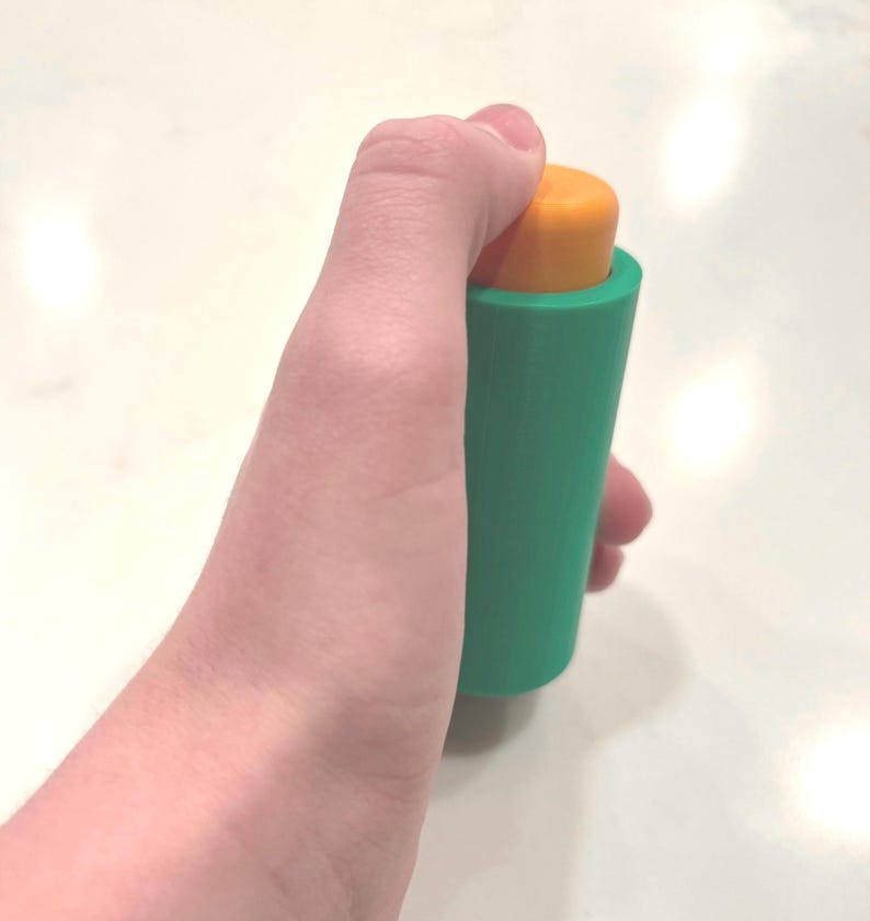 Pu&ograve; includere: Un oggetto cilindrico verde e arancione con una parte superiore arrotondata. L'oggetto &egrave; tenuto in una mano.