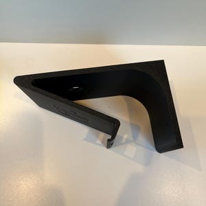 May include: Black plastic phone stand with a curved design. The stand has a small hole on the top and a cut-out on the side.