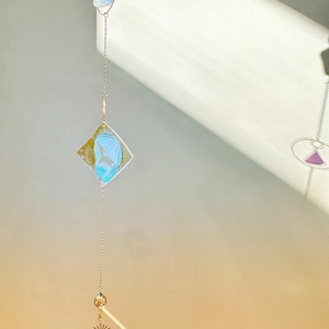 May include: A silver chain with a crystal pendant hangs from a ceiling. The pendant is a clear, faceted crystal with a gold star charm above it. A blue and gold diamond-shaped charm hangs above the crystal. A silver circle with a blue triangle charm hangs above the diamond charm.