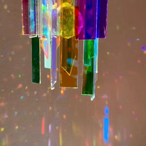 May include: A colorful wind chime with rectangular, transparent panels in shades of purple, green, yellow, and orange. A gold-colored chain and pendant hang from the top. The light refracts, creating a rainbow effect on the wall.