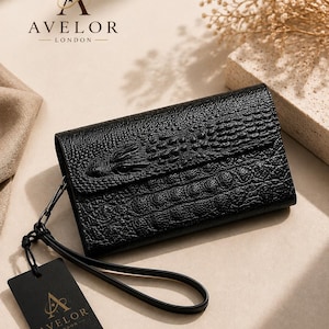 Op de afbeelding: Een zwarte rechthoekige clutch met een gestructureerd krokodillenleerpatroon. De portemonnee is voorzien van een polsbandje en een zwart rechthoekig label bevestigd aan een koord, beide met het AVELOR LONDON logo in gouden letters.