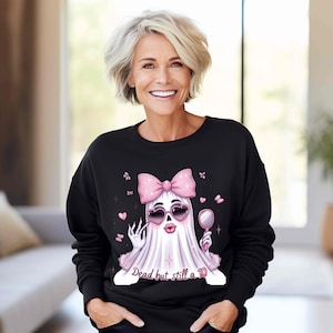 May include: A black long-sleeve sweatshirt featuring a graphic of a white ghost wearing a large pink bow, heart-shaped sunglasses, and holding a hand mirror. The ghost has pink lips and the text "Dead but still a 10" is printed below the figure.