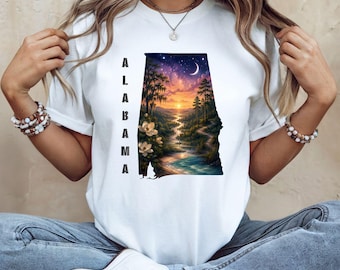 May include: A white short-sleeve t-shirt featuring a graphic of the state of Alabama. Inside the state outline is a sunset landscape with trees, a river, and flowers. The word ALABAMA is printed vertically in black capital letters to the left.