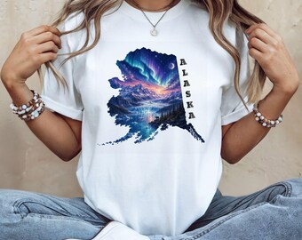 May include: A white short-sleeved t-shirt featuring a graphic shaped like the state of Alaska. The graphic displays a colorful aurora borealis over a snowy mountain landscape, a lake, and a forest. The word ALASKA is printed vertically in black.