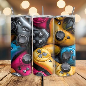 May include: Three stainless steel tumblers featuring a vibrant design of video game controllers. The controllers are in shades of blue, pink, yellow, and grey. Each tumbler has a silver straw and lid.