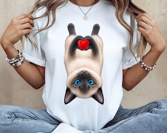 May include: A white t-shirt featuring a graphic of a Siamese cat hanging upside down. The cat has bright blue eyes and dark brown fur on its face and ears, and it is holding a small, vibrant red heart between its front paws.