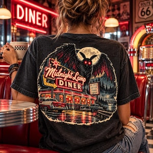 Può includere: Una t-shirt grigio antracite con una stampa grafica di un diner notturno e una grande falena sopra. Il testo recita Midnight Lamp DINER e NOT COFFEE GOOD FOOD STRANGE COMPANY. La scena include un'auto d'epoca e una luna piena.