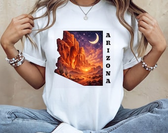 May include: A white t-shirt featuring a rectangular graphic of the state of Arizona. The design displays a desert landscape with red rock formations, cacti, a sunset, and a crescent moon in a starry sky. The word ARIZONA is printed vertically in black.