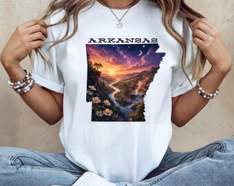 May include: A white short-sleeve t-shirt featuring a graphic print of the state of Arkansas. The design displays a colorful sunset landscape with a winding river, trees, and flowers inside the state outline, with the word ARKANSAS printed above.