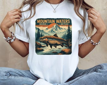 May include: A white short-sleeve t-shirt featuring a graphic print of a fish swimming in a river with mountains and a sun in the background. The text reads MOUNTAIN WATERS, WILD RIVERS, and QUIET PLACES in a dark font above the landscape illustration.