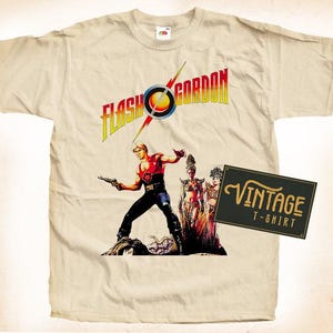 Może przedstawiać: Vintage'owa koszulka w kolorze beżowym z nadrukiem graficznym Flash Gordona, postaci z filmu z lat 80., stojącego z dwiema kobietami. Tekst "Flash Gordon" jest nadrukowany na czerwono, żółto i niebiesko.