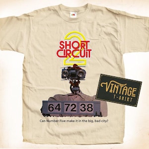 Può includere: Una maglietta beige con una grafica vintage di Short Circuit 2 che presenta Number Five, il robot, con il testo "64 72 38" e la domanda "Can Number Five make it in the big, bad city?" La maglietta ha anche un'etichetta di maglietta vintage.