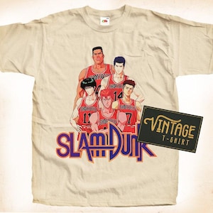 Può includere: Una maglietta vintage con un gruppo di giocatori di basket in uniforme rosso e bianco. I giocatori sorridono tutti e tengono in mano palloni da basket. Il testo "SLAM DUNK" è stampato in viola e giallo sulla parte anteriore della maglietta.