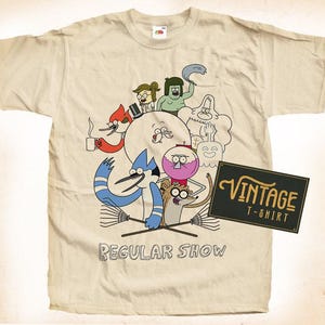 May include: Beige vintage t-shirt featuring a colorful graphic of characters from the animated series "Regular Show." The design includes a large white circle with several cartoon figures, the text "REGULAR SHOW" and a black sign that says "VINTAGE T-SHIRT."