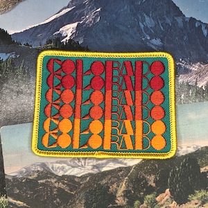 May include: A square-shaped embroidered patch with a teal background and a yellow border. The patch features the word "Colorado" repeated multiple times in a retro font with orange, red, and yellow colors.
