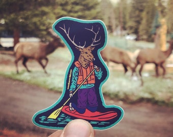 May include: A sticker of an elk wearing a life vest and holding a paddle, standing on a red surfboard in blue water. The sticker is on a white background and has the text "woodenswift.com" on the side.