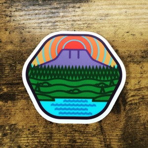 May include: A colorful sticker with a stylized illustration of a mountain, trees, and a lake. The mountain is purple, the trees are green, and the lake is blue. The sun is setting behind the mountain, and the sky is orange and yellow.
