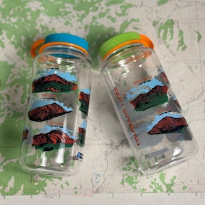 Puede incluir: Dos botellas de agua de plástico transparente con gráficos de montañas coloridas. Una tiene una tapa azul y un asa naranja, la otra tiene una tapa verde y un asa naranja. Las botellas están sobre un mapa.
